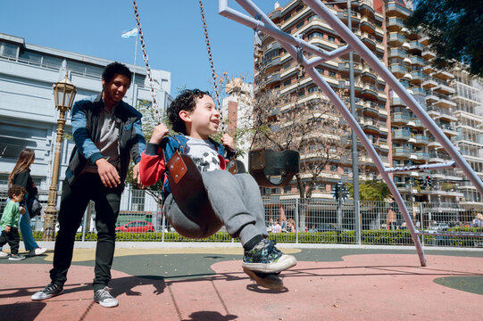 Happy Young Father Laughing And Playing With His Little Male Child Pushing Pain On Swing In Park