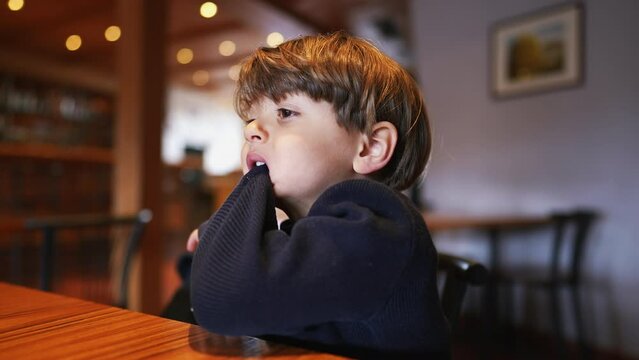 Bored small boy pulling sleeve with teeth and mouth while seated at restaurant with nothing to do. Child bites sweater feeling boredom
