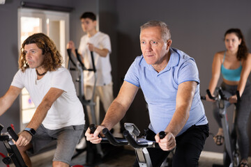 Fototapeta na wymiar Portrait of elderly man taking indoor cycling class at fitness center, doing cardio riding bike