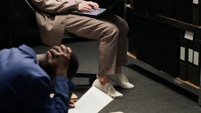 African american male officer and caucasian female inspector with laptop analyze evidence in incident room. Tired policewoman sits on chair while policeman on the floor examining clues and statements.