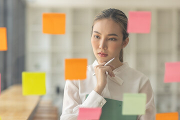 business asian girl in white shirt thinks, holds pen and clipboard, writing sticking adhesive notes on glass wall, making notes, standing over white background