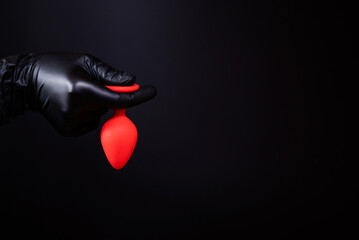 Female hand in a black latex glove holds a red anal plug on a black isolated background. Sex toys,...