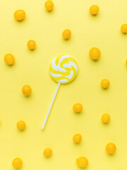 A yellow lollipop on a yellow background with scattered yellow candies. The concept of sweets.
