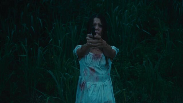 A frightened girl in a bloodstained dress stands with a gun in her hands aiming at her pursuer