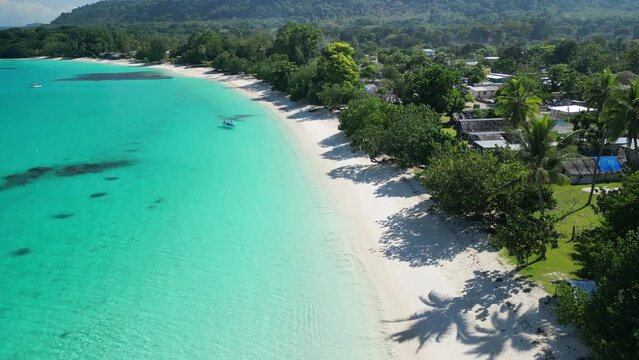 A drone flies over a stunning white sand beach with a red boat at Port Olry on the island of Espiritu Santo. The untouched area in remote Vanuatu is known for its green hills and stunning blue water.
