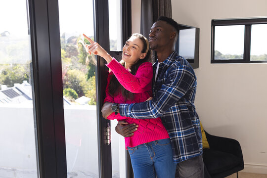 Happy diverse couple embracing and looking through window at home
