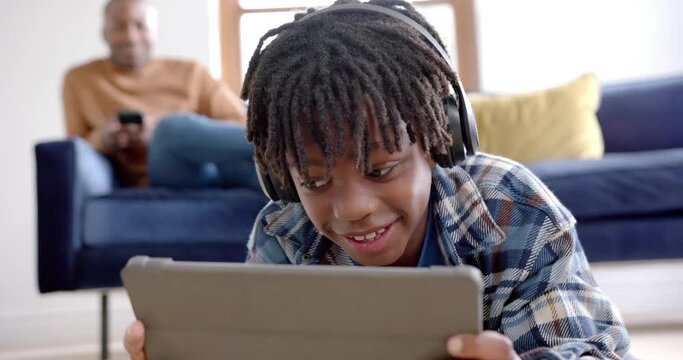 Happy african american boy with headphones using tablet lying on floor at home, slow motion
