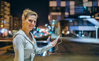 Portrait of smiling young blonde sportswoman listening music on mobile phone application. Female runner with earphones looking at camera after training at night on empty town. Right copy space.