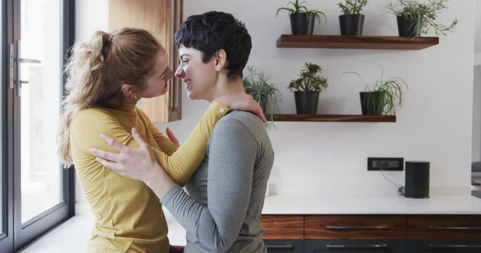 Happy caucasian lesbian couple embracing and kissing in sunny kitchen, copy space