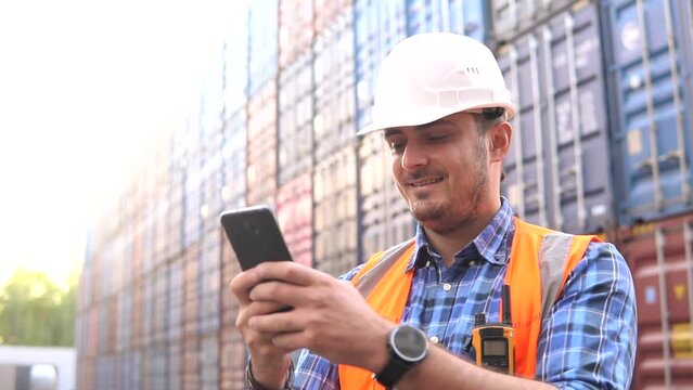 Young man engineer in the hard hat using phone in container warehouse. Logistics management in container yard.
