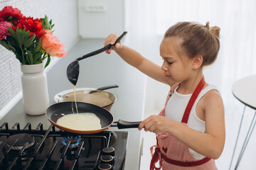A little 8-year-old girl in an apron is frying pancakes in the kitchen.
