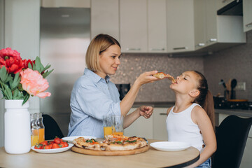 Mom and daughter are eating delicious pizza in the kitchen.