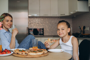 Mom and daughter are eating delicious pizza in the kitchen.