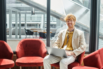 cheerful red haired man with laptop on his laps sitting on chair with glass backdrop, coworking