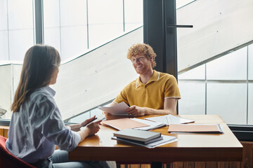 happy coworkers sitting at table and smiling at each other holding phone and work papers, coworking