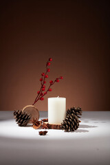 Minimal scene of a white scented candle displayed with few pine cones and star anise. Brown...