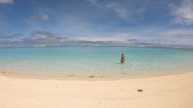 Still view of turquoise clear sea salt water Ocean Beach at shore with white sand over look out to horizon with blonde Caucasian young woman girl dressed in black bikini in Aroa Beach Raro