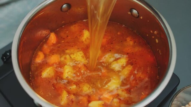 Pouring the broth into a pan with fried vegetables with spices. Close-up of the table. Cooking in the kitchen