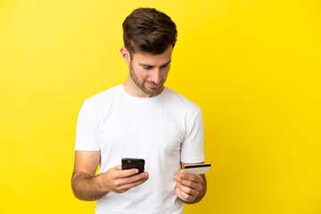 Young caucasian handsome man isolated on yellow background buying with the mobile with a credit card