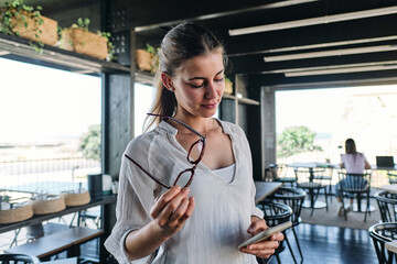 Portrait of pretty blonde standing in cafe with eyeglasses and smart phone in hands