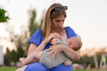 young white mother in blue dress sitting outdoors and breastfeeding her newborn daughter