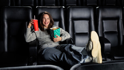 A laughing young woman is watching a comedy at the cinema. A woman drinks soda and eats popcorn