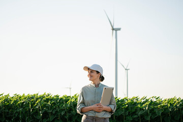 Smiling agronomist holding laptop at field with wind turbines in background