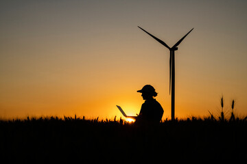 Silhouette of farmer using laptop in field with wind turbine at dusk