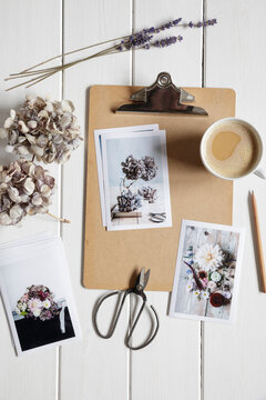 Cup of coffee, clipboard, dried flowers and photographs of flower arrangements lying on white painted wood