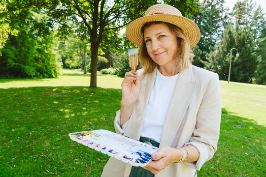 Smiling mature woman wearing hat holding paintbrush and watercolors