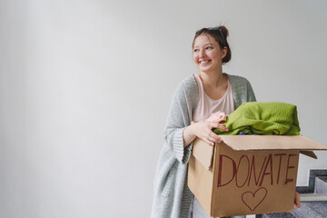 Smiling teenage girl holding box with clothes to donate in front of wall