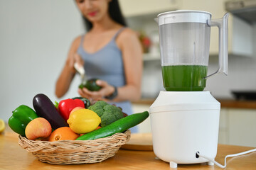 Blender and fresh vegetables on kitchen countertop with young woman cooking in background. Dieting, healthy food concept
