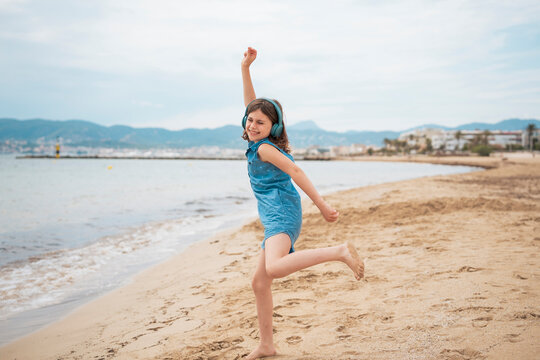 Cheerful girl wearing wireless headphones listening to music and dancing on coastline at beach