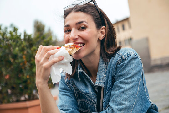 Smiling woman eating pizza sitting on road