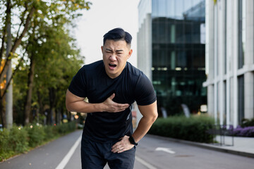 Asian young male sportsman stands bent over on the city street and holds his hand to his chest, felt severe pain and spasm in his heart