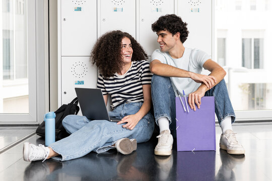 Young students sitting with laptop and laughing in front of locker in university