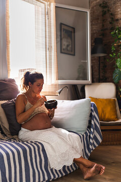 Pregnant woman eating oatmeal on sofa at home
