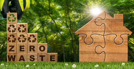Gloved hand arranging wood blocks with text Zero Waste and Recycling Symbols on a green meadow with a small wooden house made of jigsaw puzzle pieces. Green forest on background.