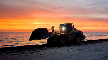Bulldozer on the beach at sunset. A large tractor cleans the beach of sea grass. Bulldozer in motion and blur. - obrazy, fototapety, plakaty