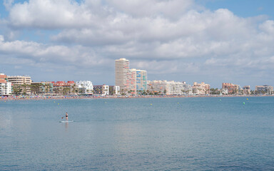 Fototapeta premium Panoramic image of Peñíscola (Spain), a crowded Spanish beach, with buildings, many people on vacation, tourists bathing and paddle surfing, under the summer heat.