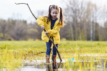 Girl holding branch while standing in puddle and looking down with focused eyes, preparing for game with paper boats.