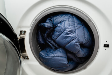 Blue puffer jacket in the drum of open washing machine in laundry room. Washing dirty down jacket in the washer - obrazy, fototapety, plakaty