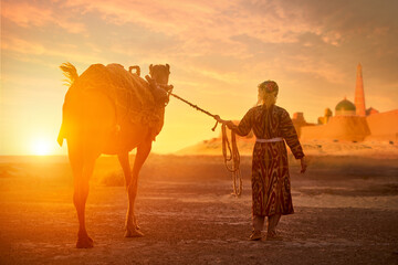 Woman in traditional national clothing leads camel through desert towards ancient city of Khiva at sunset. - 657007968