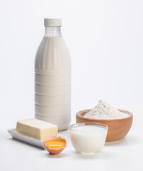 fresh milk in plastic bottle with white flour in wooden bowl, yogurt in glass bowl, broken egg and butter on white plate all on white background