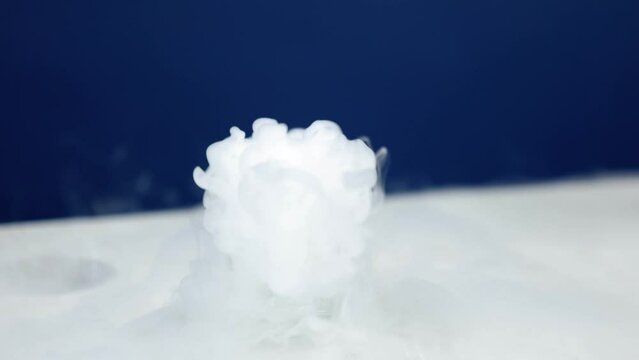 A beaker in a laboratory emits a cascade of white smoke, bubbling and swirling mysteriously against a blue background.