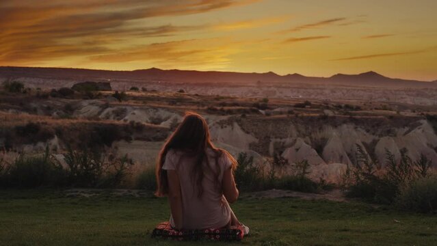 A young woman has come to enjoy the sunset. She laid a pillow on the grass and sat down, with a view of the mountains and the canyon in front of her.