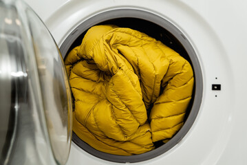 Yellow winter puffer jacket in the drum of open washing machine in laundry room. Washing dirty down jacket in the washer - obrazy, fototapety, plakaty