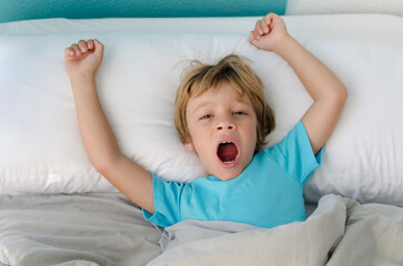 blond boy yawning in the morning waking up in bed in a very bright room. 6 year old child looking...