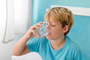 Pre-teen boy starts his morning with a glass of fresh water in bed. Handsome kid of ten years old drinking water sitting on the bed at home in the morning.  Healthy lifestyle concept
