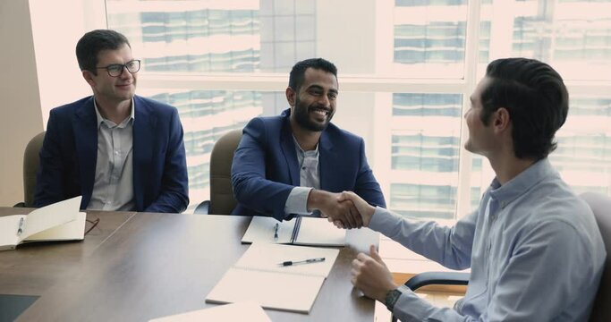Three multi-ethnic businessmen negotiate together in boardroom, make profitable commercial deal, shake hands express respect after effective formal meeting. Trust, legal commitment, business etiquette